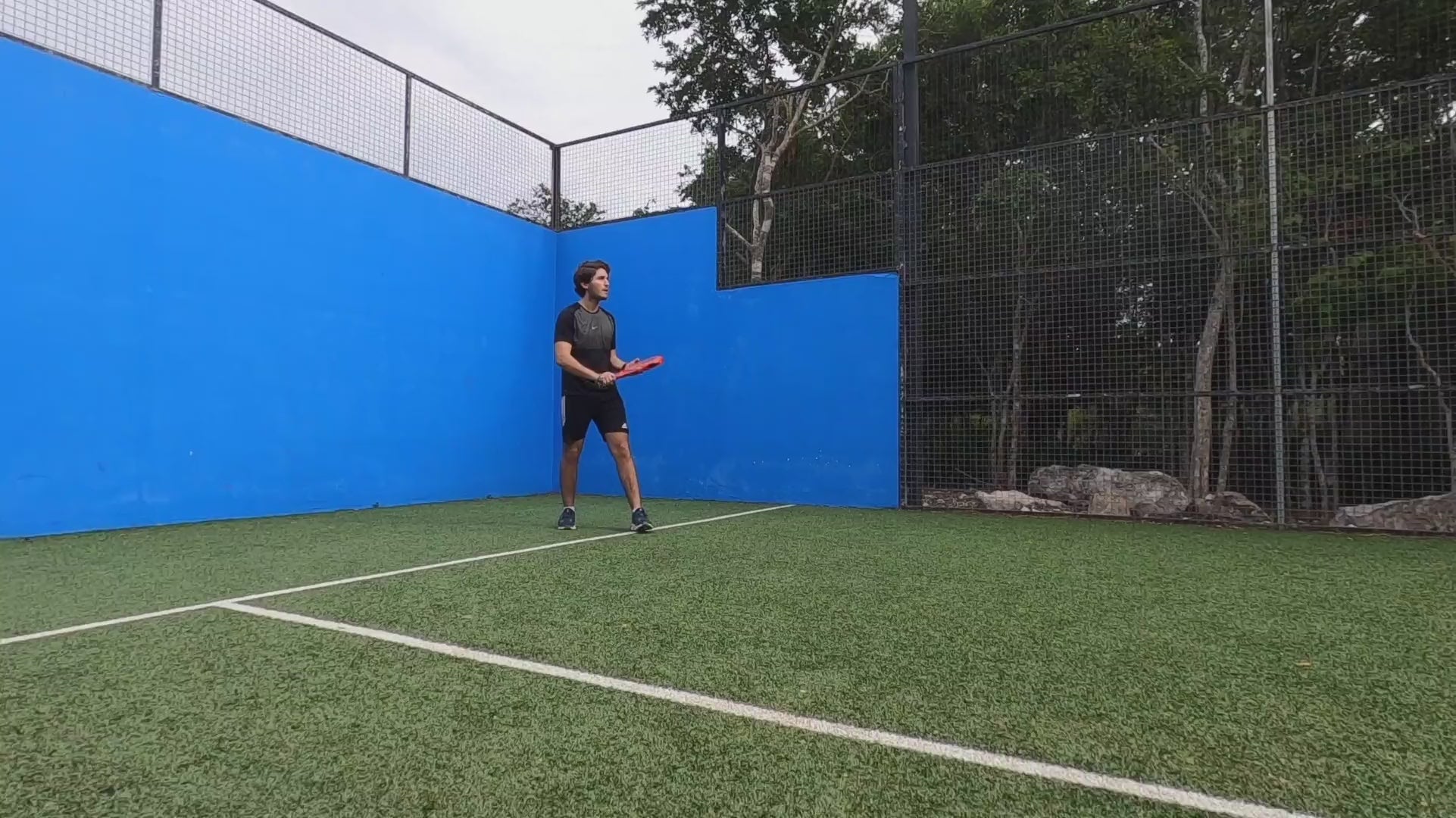 Jeune joueur réalisant un coup droit avec une raquette rouge sur un terrain de padel extérieur aux murs bleus.